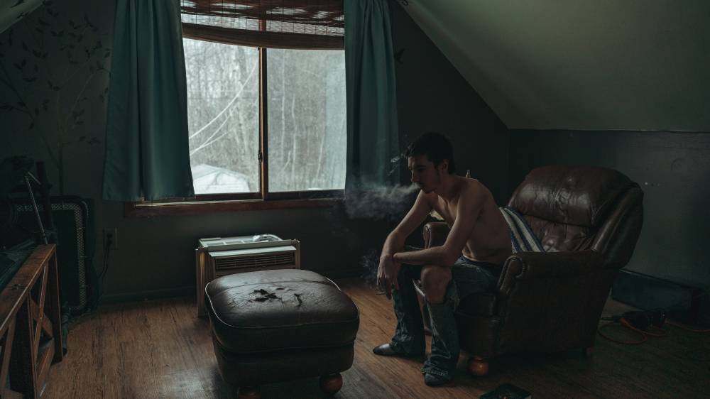 Young shirtless man sitting in darkened room smoking a cigarette